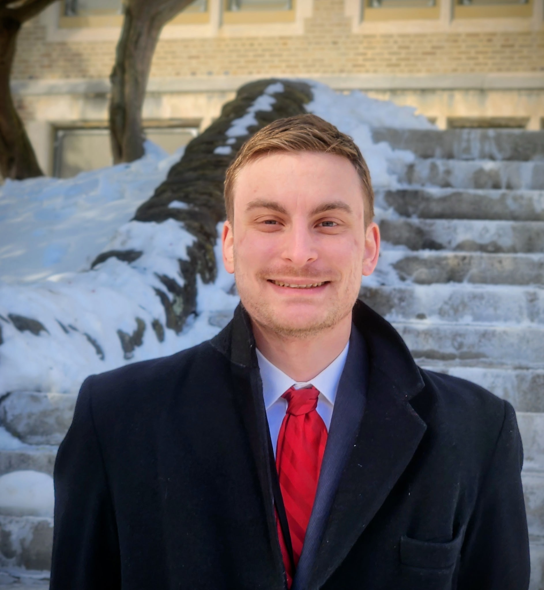 Portrait of Alex Schnell in a suit and red tie standing in front of stone steps